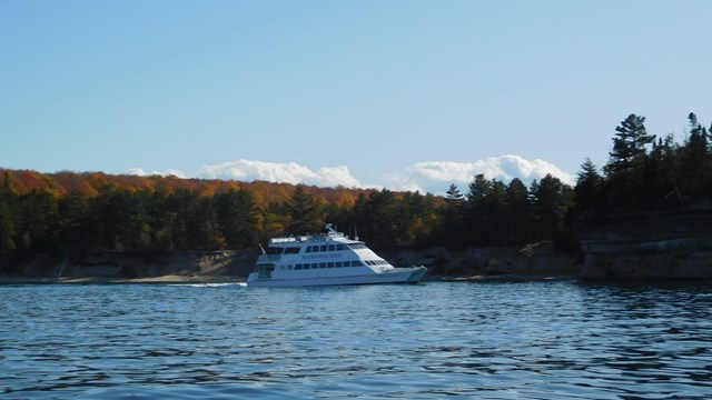A white cruise ship sits in the water in front of autumn foliage.