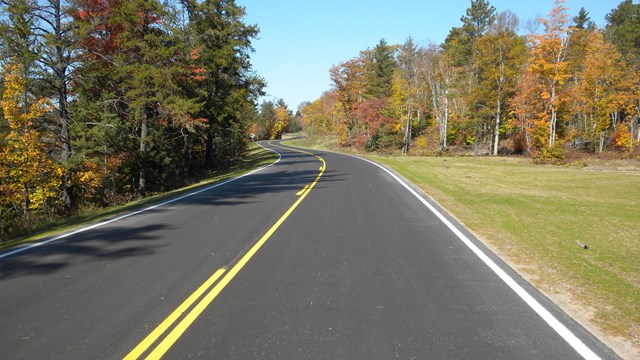 A blacktop road with yellow lines extends in front of the viewer. Fall foliage is on either side
