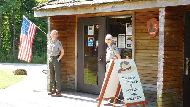 Two rangers stand in front of a visitor center with an American flag. Trees are in the background. 