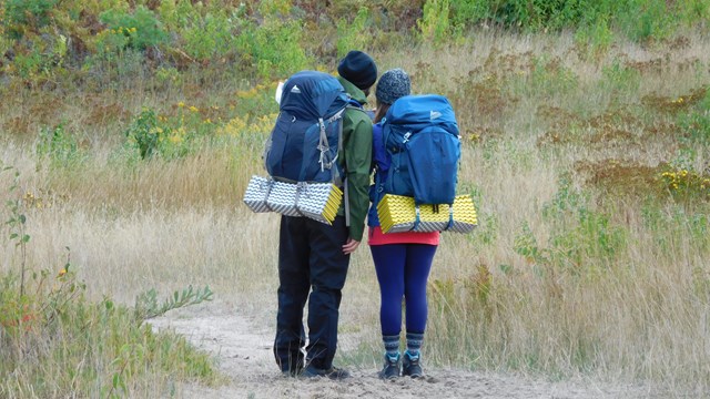 Two people wearing large backpacks look at a map at a trailhead. 