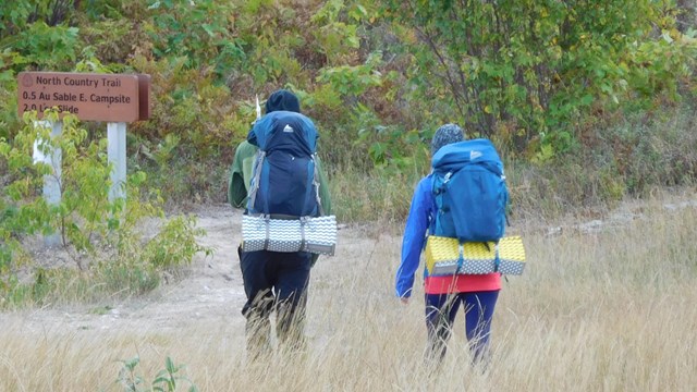 Two people wearing large backpacks walk away from the camera towards a trail.