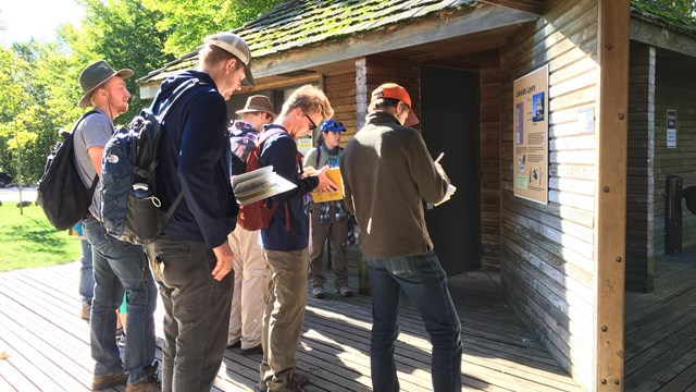 A group of people wearing hats and backpacks looking at books and notes while in front of a sign.