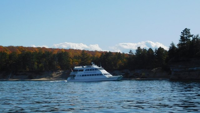 A white cruise ship sits in the water in front of trees in autumn colors.