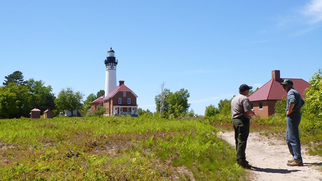 Two people stand in front of a field with a large white lighthouse in the background.