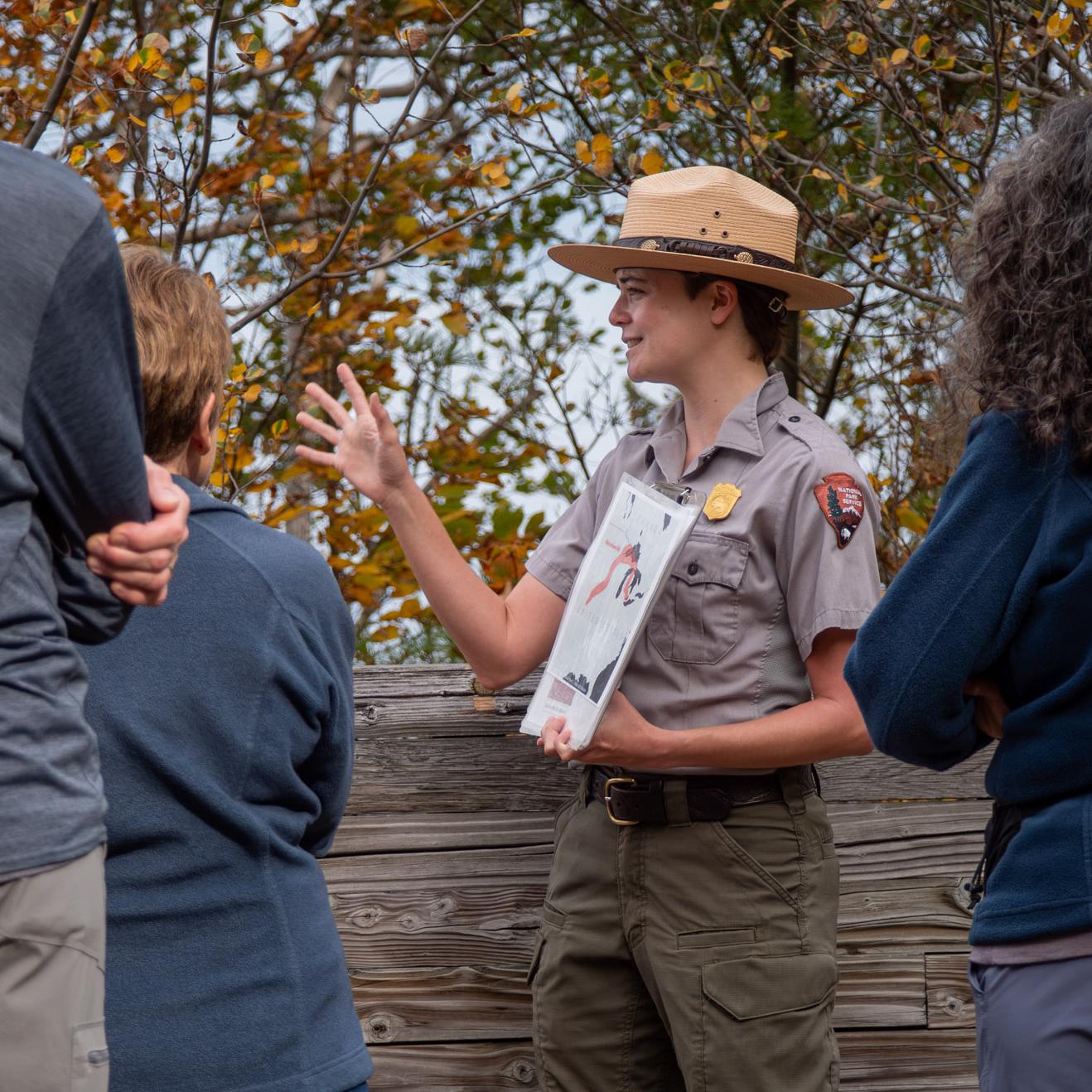 A ranger standing in front of a wooden wall and autumn leaves talks to a group of people.