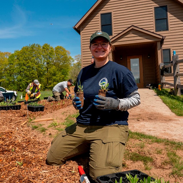 A person working in a garden smiles while holding two plant plugs