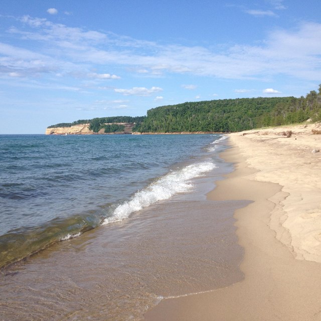 Small waves land on Miners Beach. With trees and cliffs in distant.