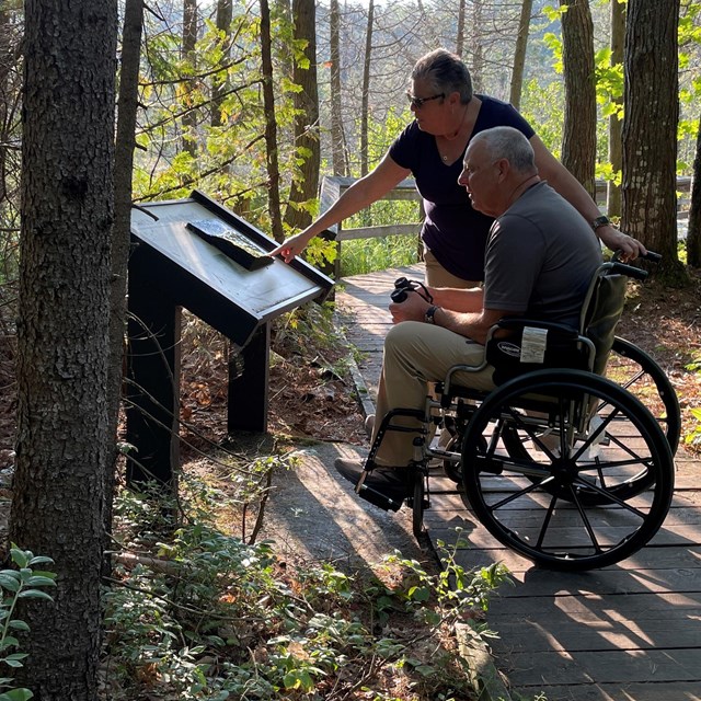 Two visitors on boardwalk trail, one in wheelchair, looking at a wayside exhibit.