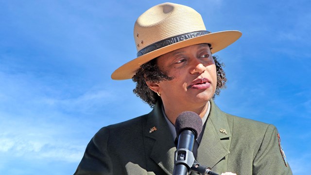 An African American woman in a ranger uniform stands in front of a microphone. She is outdoors. 