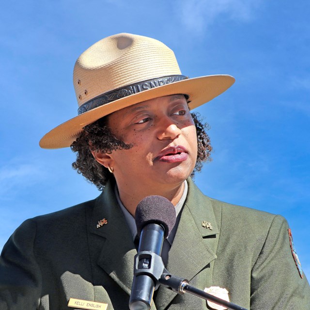 An African American woman in a ranger uniform stands in front of a microphone. She is outdoors. 