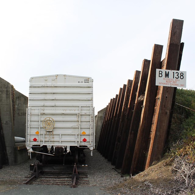 A box car sits on a rail track, between two large wooded walls, backed by earth mounds. 