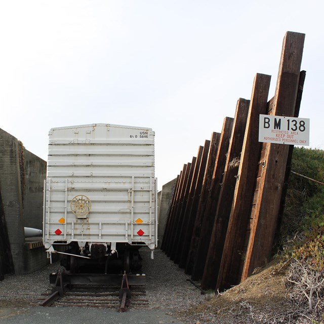 A box car sits on a rail track between two large wooded walls, backed by earth mounds. 