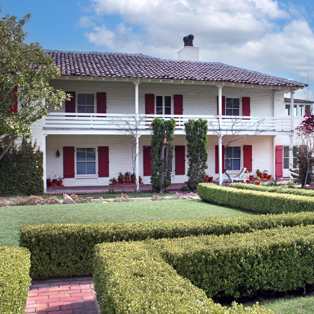 A two-story home with an upper balcony and shutters. A courtyard full of trees, shrubs and plants. 