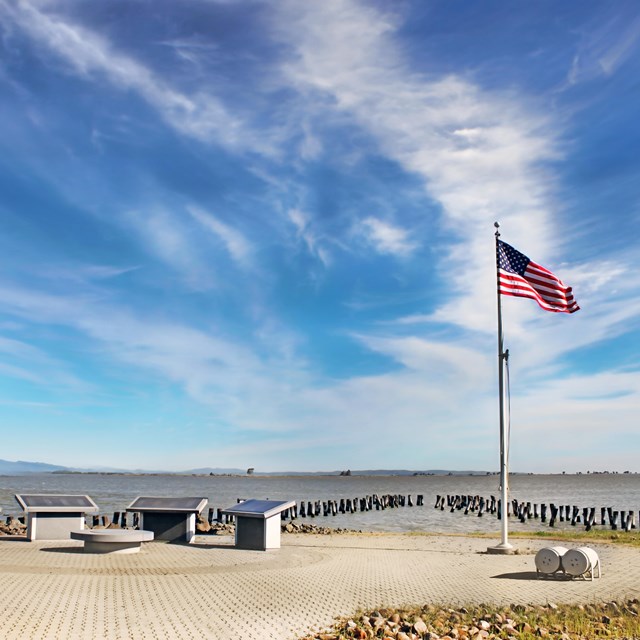 Four memorial stones sit on layered brick. An American flag waves in the wind. Bay water and hills. 