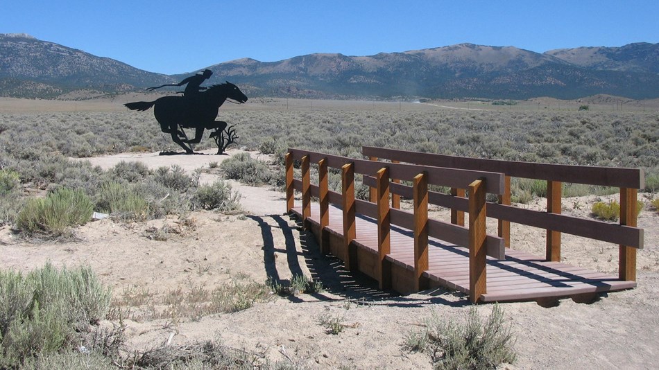 A black steel silhouette of a Pony Express rider behind a wooden footbridge with mountains.
