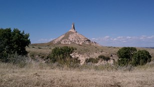 A pointed bluff landmark sticks out above a flat valley with large green shrubs.