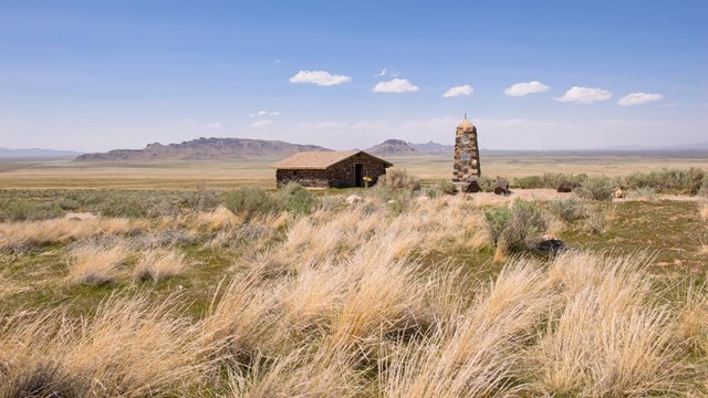 Tall grass leads to a distant barn set in an expansive grassland with distant mountains.