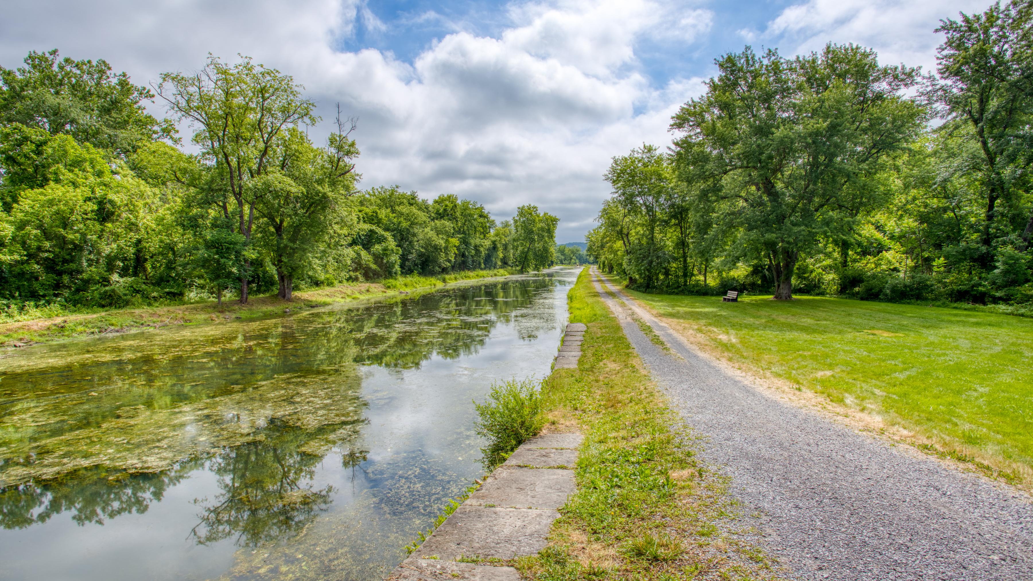 A body of water in a canal next to a gravel path. The trees are on both sides and the sky is cloudy.