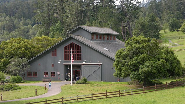 Visitors at the entrance of a gray barn-like visitor center surrounded by a fenced green pasture.
