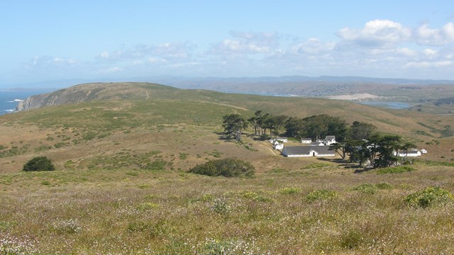 A ranch complex with white wooden buildings on a narrow peninsula between the ocean and a bay.