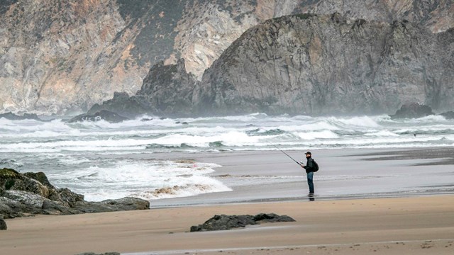 A man fishing on a sandy beach with waves washing in from the left & steep bluffs in the background.