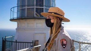 A females ranger wearing a face mask stands in front of a lighthouse.