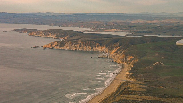 An aerial photo of a long green peninsula of land, with steep cliffsides and sandy beaches.