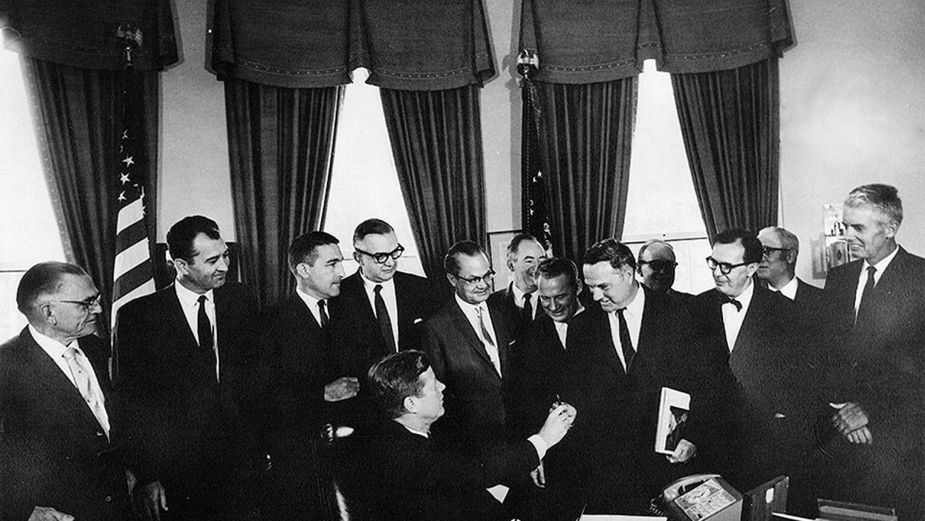 Eleven men in suits look on as a man seated at a presidential desk hands his pen to the man behind.