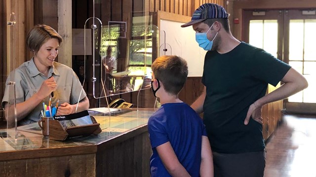A park volunteer with blond hair talks to a father and son at a visitor center desk.