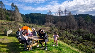 A family of seven sits around a picnic table in a campsite with a tent and food storage locker.