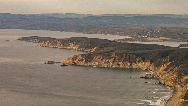 An aerial photo of a long green peninsula of land, with steep cliffsides and sandy beaches.