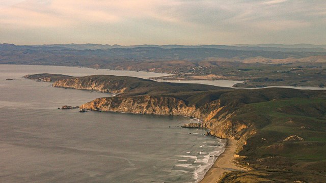 An aerial photo of a long green peninsula of land, with steep cliffsides and sandy beaches.