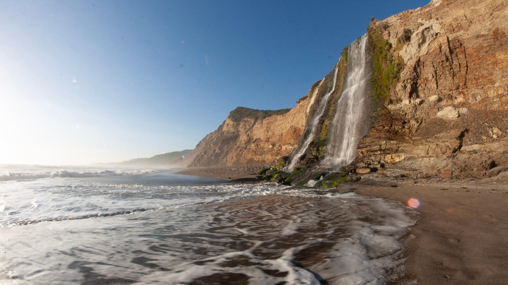A 40-foot-tall waterfall cascades over coastal bluffs on the right onto a sandy ocean beach.