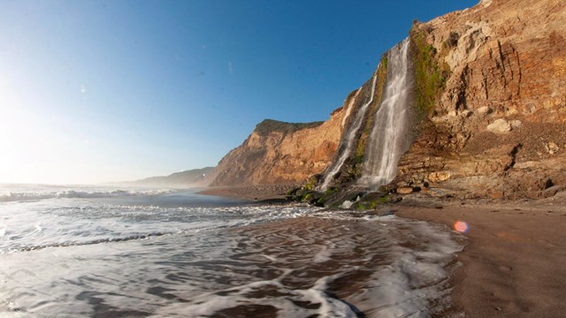 A 40-foot-tall waterfall cascades over coastal bluffs on the right onto a sandy ocean beach.