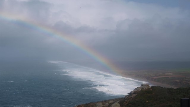 A rainbow arcs up and to the left from a beach on the right over the ocean through some clouds.