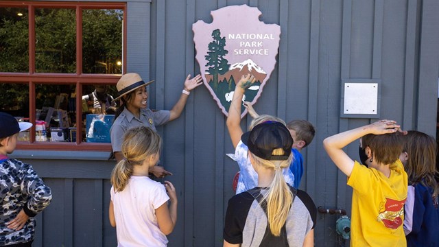 A female ranger gesturing toward a 3-foot-tall wooden NPS arrowhead talks with several children.