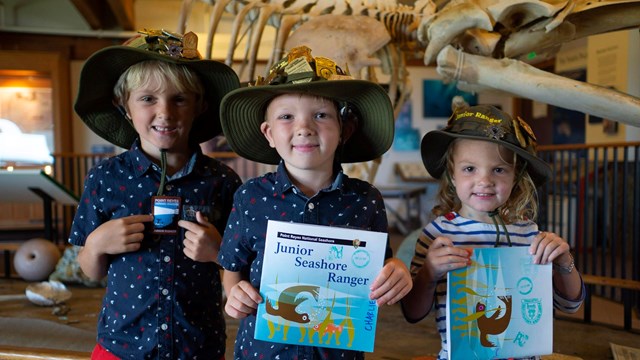 Three young junior rangers pose with their booklets, badges, & patches in front of a whale skeleton.