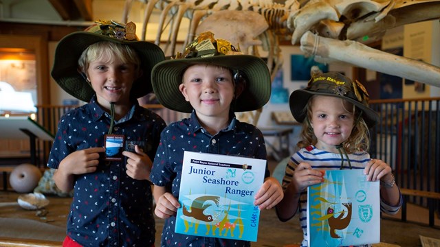 Three young junior rangers pose with their booklets, badges, & patches in front of a whale skeleton.