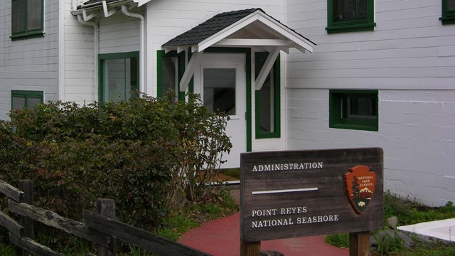 A photo of a doorway of a white two-story building beyond a brown wooden "Administration" sign.