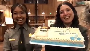 Two smiling, female rangers in a visitor center hold a layer cake decorated with a lighthouse.