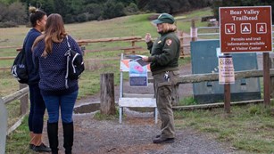 A female ranger talks with two female hikers at a trailhead. Signs indicate the trail is closed.