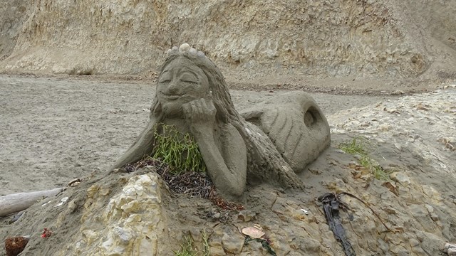 A medium-sized sand sculpture of a mermaid lying on her belly on top of a rock outcrop.