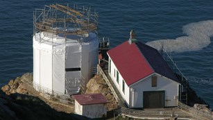 White structures, some with red roofs, surrounded by scaffolding perched on a cliff above the ocean.