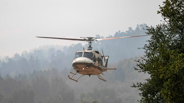 A white helicopter flies over a smoky, treed ridgeline. 