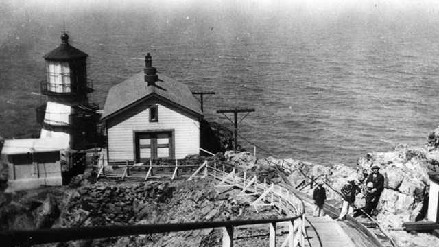 Black and white photograph of concrete stairs leading down to a cliffside lighthouse.