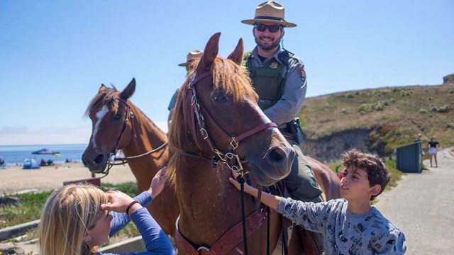 Two kids petting a horse atop which is a smiling park ranger.