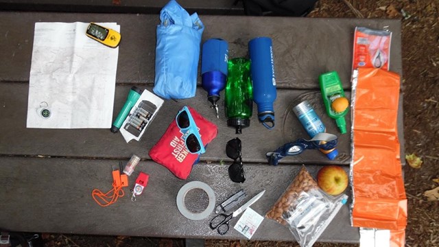 Essentials for hiking spread out over a picnic table: a flashlight, a map, sunglasses, food, etc.