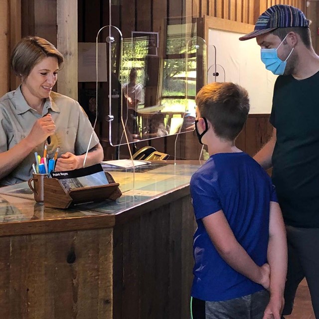 A woman wearing a khaki-colored shirt stands behind an information desk while assisting a visitors.