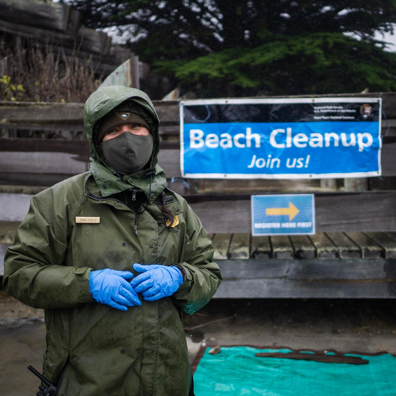 A park ranger in a hooded green coat stands in the rain in front of a blue banner. 