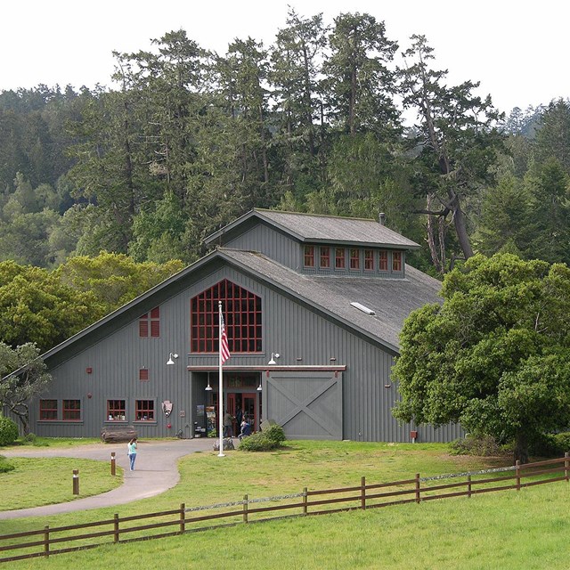 Visitors at the entrance of a gray barn-like visitor center surrounded by a fenced green pasture.